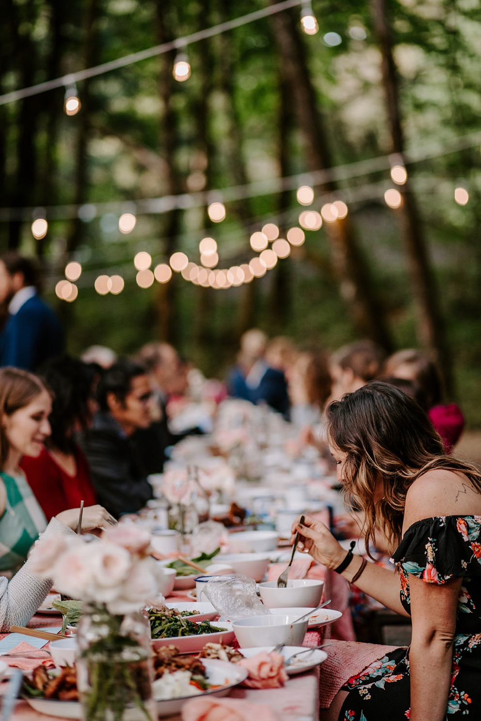 guests seated at long table in forest under patio lights during outdoor wedding reception
