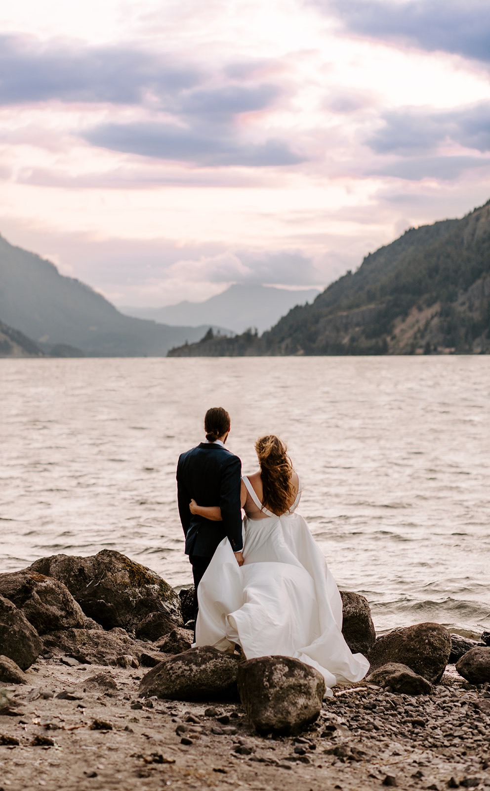 bride and groom look across water toward mountains as sun sets