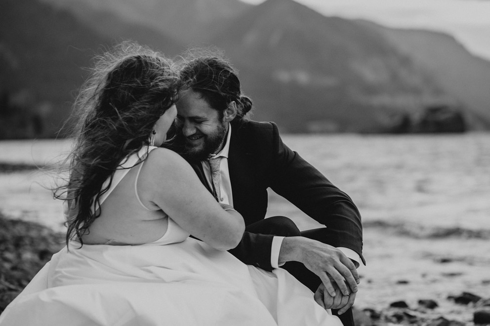 bride and groom sit smiling near water's edge