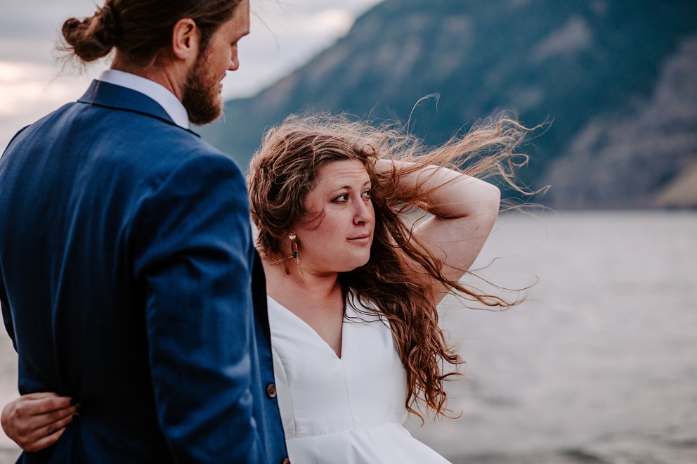 bride stands near water's edge in the wind